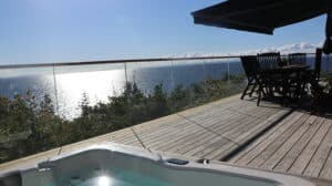 Stolpefrit glasværn på terrasse ved havet med trægulv og havemøbler i skygge under markise.