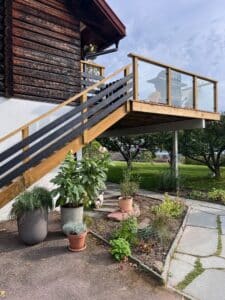 Højtliggende altan med rækværk i træ og klart glas ved bjælkehytte, med udsigt over have og grønne omgivelser.