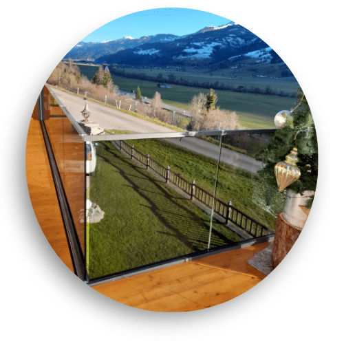 Stilrent stolpefrit glasværn monteret på træterrasse med panoramaudsigt over åbent landskab og snedækkede bjerge.