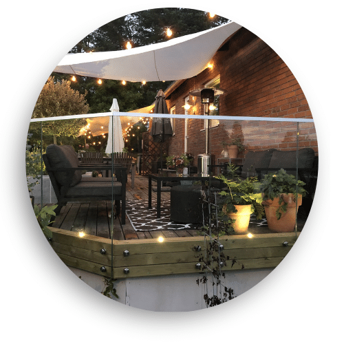 Træterrasse med helglasværn, omgivet af havemøbler, planter og stemningsfuld udendørsbelysning.