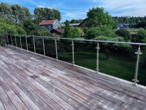 Altan med glashegn i røgfarvet sikkerhedsglas og rustfrit stål, med udsigt til have og bolig.