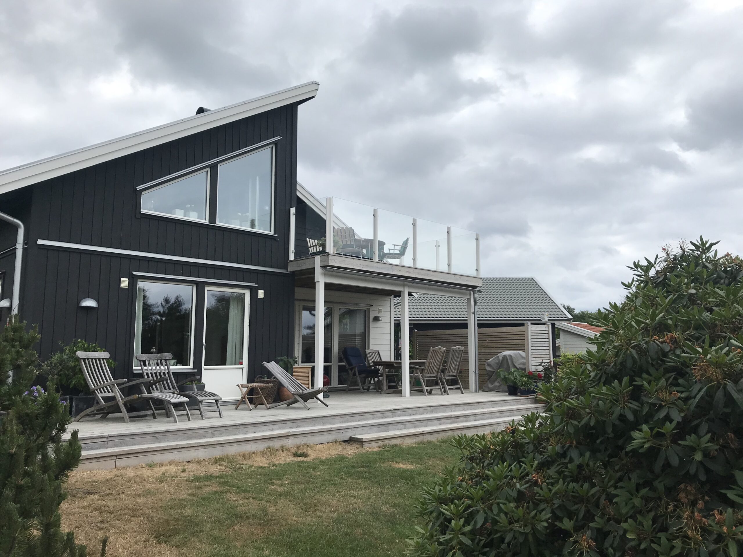 Klart glasgelænder monteret med klemmer på træterrasse ved sort træhus med skråtag og store vinduer
