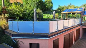 Terrasse med frostet glasværn og runde stolper i rustfrit stål, omgivet af grøn have og udestue.