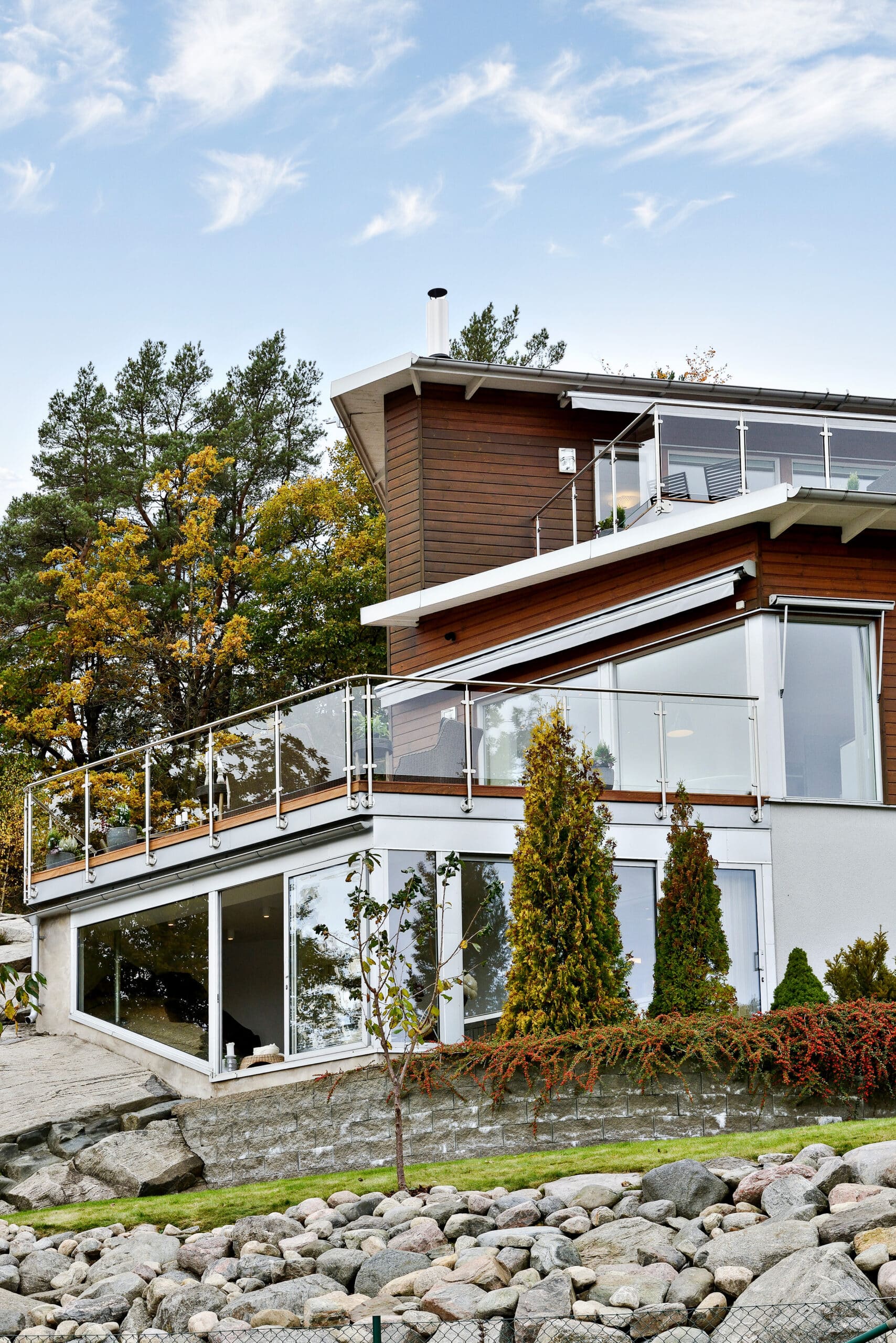 Slankt rustfrit gelænder med glas omkring hævede terrasser med natursten og høje graner