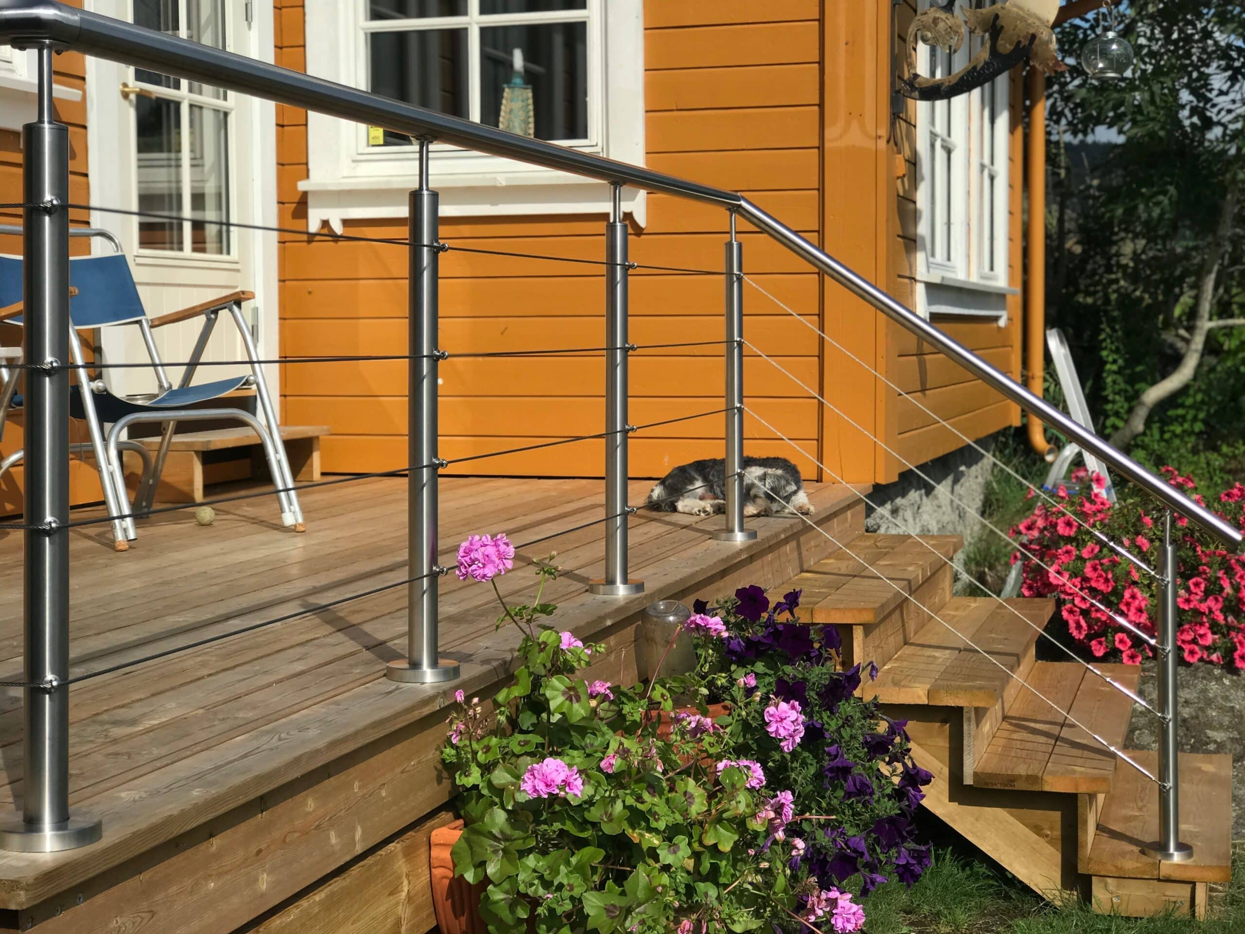 Smukt vajerrækværk i rustfrit stål med skrå sektioner langs trætrappe, blomsterkrukker og altan foran gul træfacade.