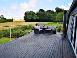 Altan med rækværk i rustfrit stål og wirer, grå terrassebrædder og siddeområde med udsigt over mark og landskab.