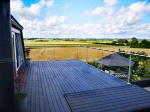 Rækværk i rustfrit stål og wiresystem på altan med mørkegrå trædæk, med frit udsyn over marker og grønne enge.