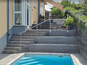 Rustfrit gelænder ved trappe og pool Gelænder i rustfrit stål monteret ved bred trætrappe ved pool – supplerer glashegn og giver komplet udendørs afskærmning.