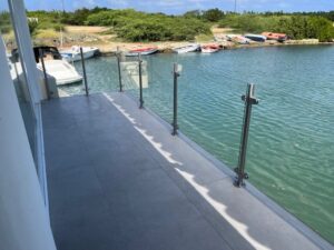 Glasværn med rustfrie stolper langs bro med udsigt til småbåde og grøn baggrund.