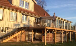 Glasværn i rustfrit stål og klart glas monteret langs trappe og terrasse i træ med udestue og solrig facade.