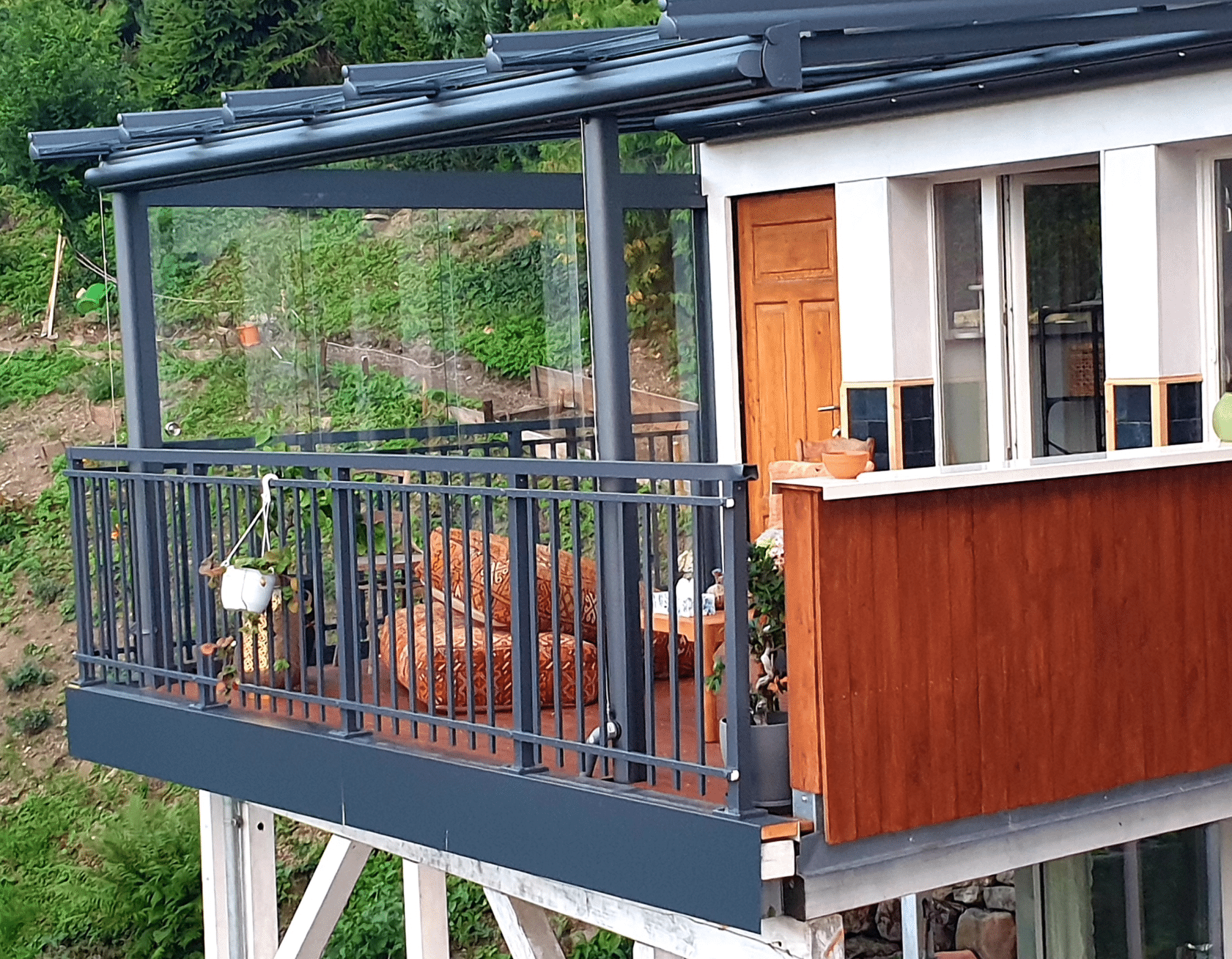 Sort aluminiumsgelænder i smedestil langs glasinddækket terrasse på skrånende hus med træfacade