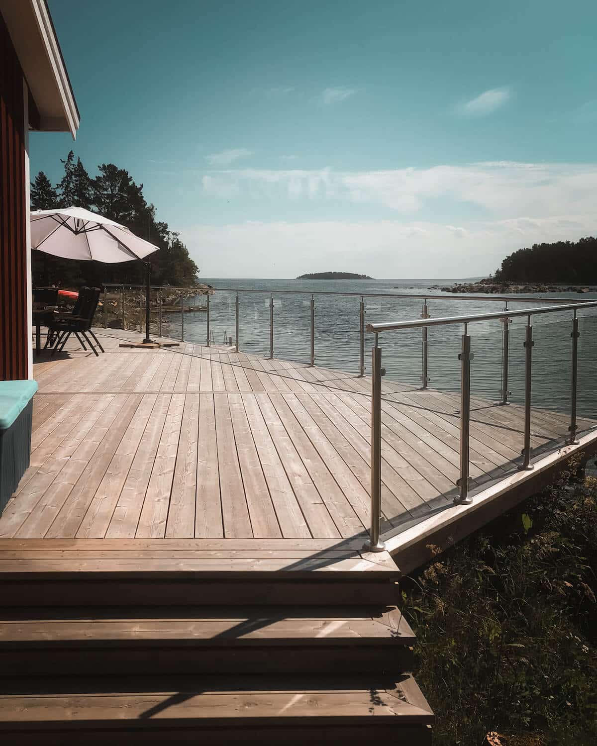 Slankt rustfrit og glasgelænder omkring stor træterrasse med havudsigt og blå himmel