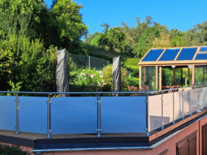 Balkonhegn i rustfrit stål med frostet glas, monteret på en hævet terrasse. Runde stolper og overligger i metal. Baggrunden viser en frodig have, drivhus med glastag og blå himmel.