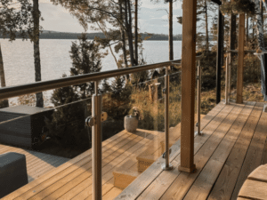 Glashegn med rustfri stolper og træoverligger monteret på træterrasse. Klembeslag fastholder glaspanelerne. I baggrunden ses en skovsø og en trappe i træ, der fører ned til vandet.